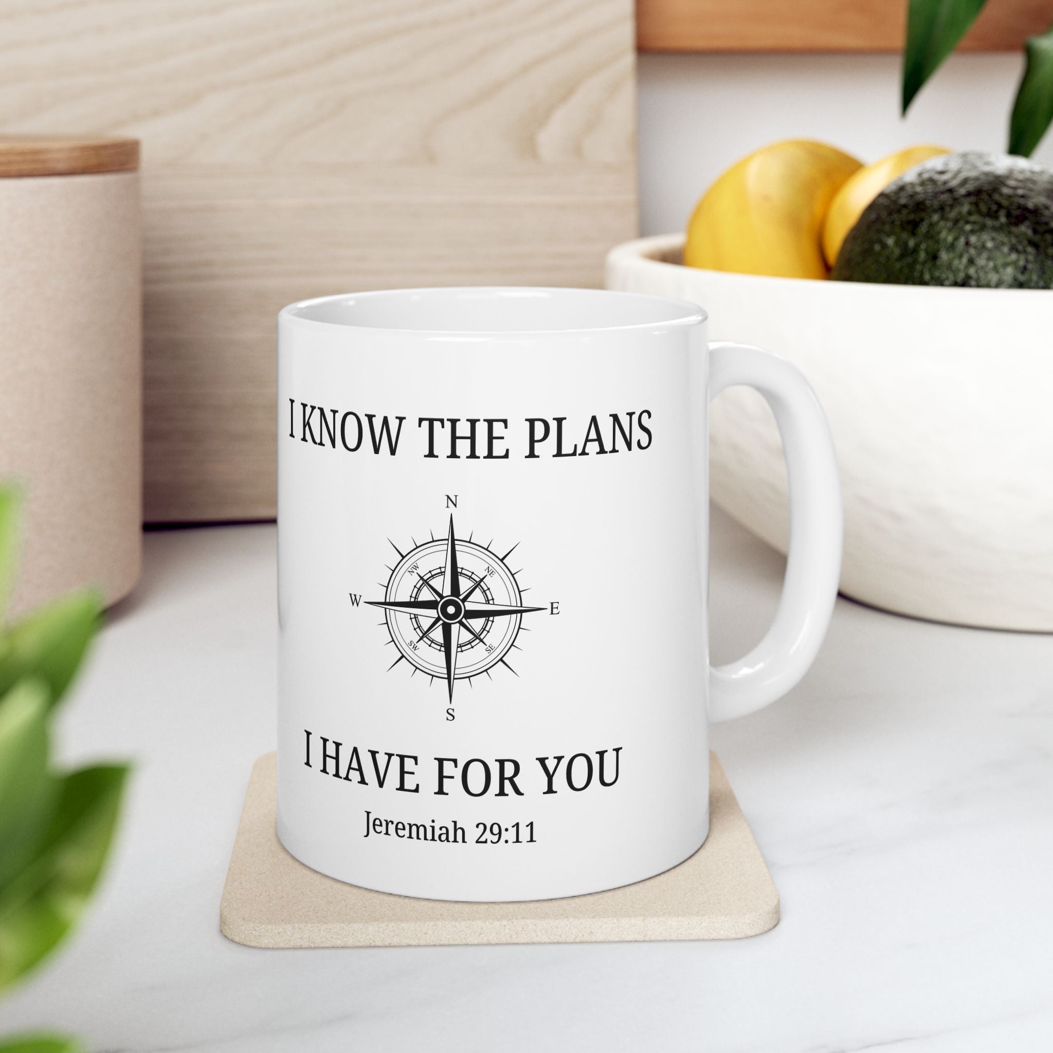 White mug with motivational quote and compass design on a coaster, placed on a surface with plants and containers in the background.