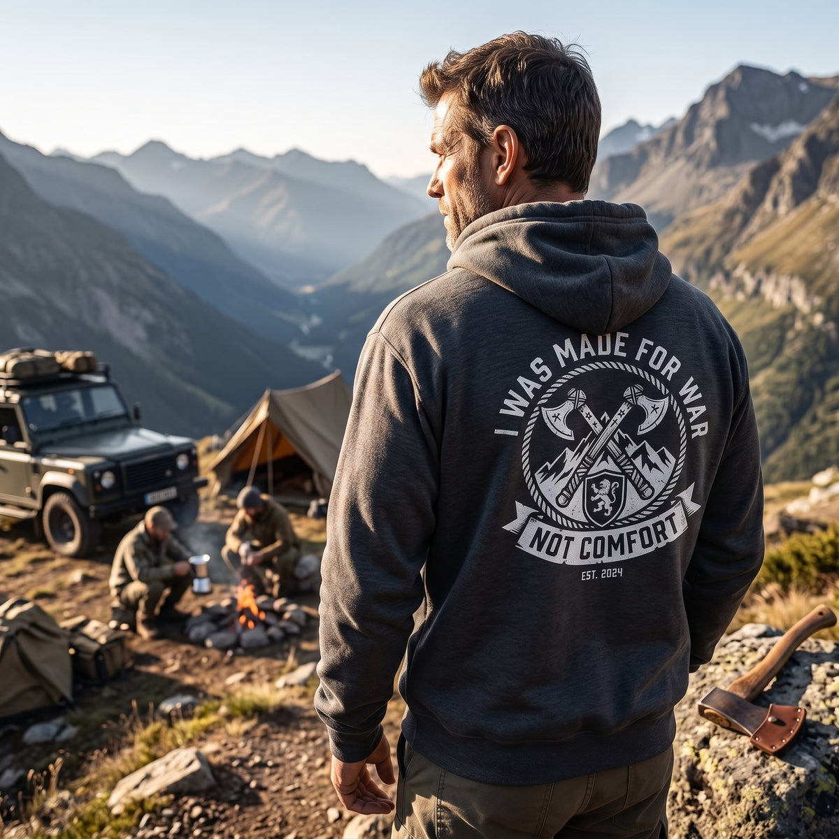 Man wearing I Was Made for War Not Comfort faith warrior hoodie in dark heather, standing on mountain summit overlooking alpine camp with Land Rover and campfire