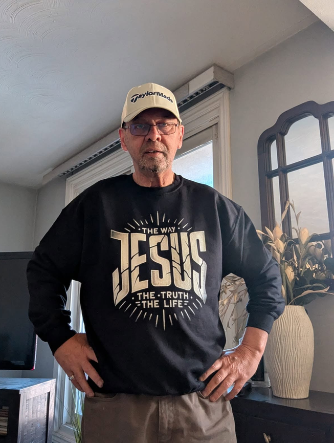 A man wearing a black long-sleeve crewneck sweatshirt with a large cream graphic that reads 'THE WAY JESUS THE-TRUTH - THE LIFE' and a cream TaylorMade baseball cap, standing in a home interior.