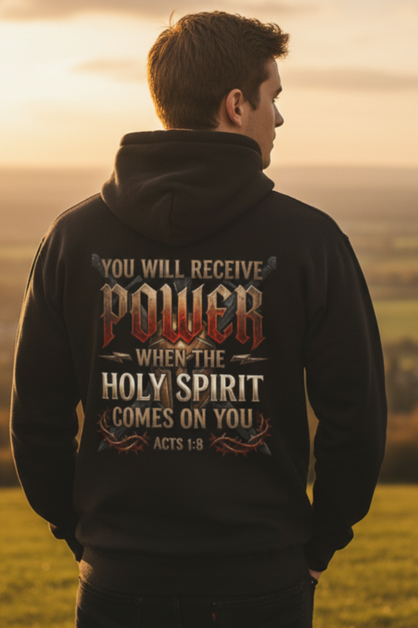 Young man wearing a black Acts 1:8 "You Will Receive Power When the Holy Spirit Comes On You" Christian hoodie, photographed from behind while standing in an open field under a warm golden sunset sky.