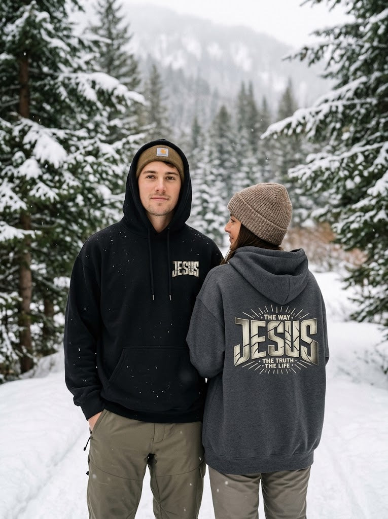 Couple on a snowy forest trail wearing Jesus The Way The Truth The Life hoodies: man in black with JESUS on the chest, woman in dark heather gray showing the John 14:6 back design